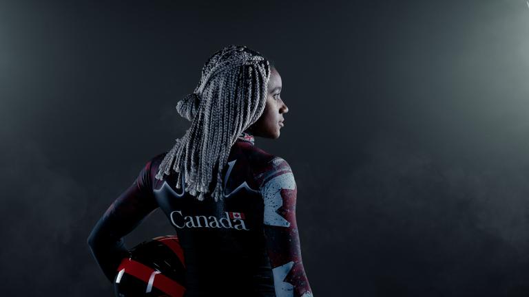 Une femme noire se tient dans un endroit sombre entre deux grands projecteurs. Elle tient un casque et porte une combinaison de bobsleigh canadienne. Visibilité masquée.