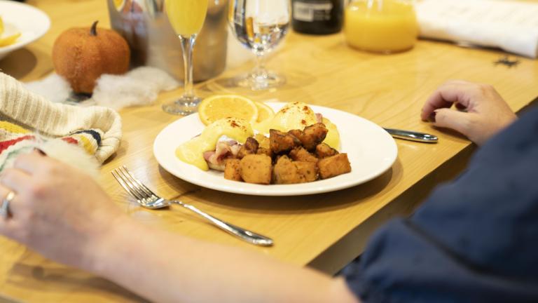 Une personne est assise à une table avec un verre de jus d’orange et une assiette d’œufs bénédictine et de pommes de terre rissolées. Visibilité masquée.