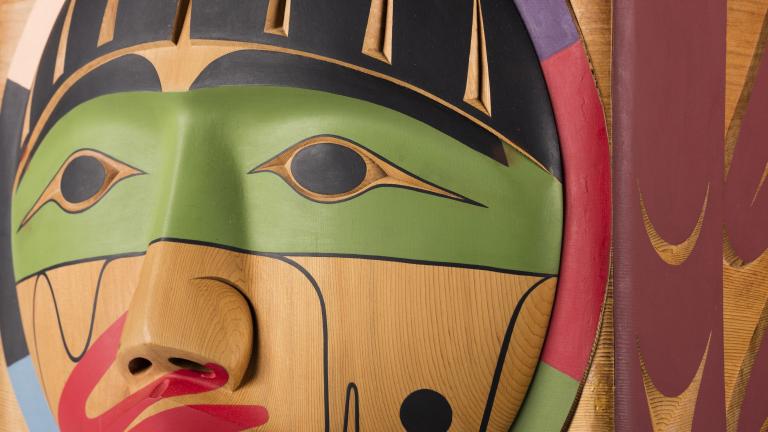 Un plan rapproché d’une boîte en bois, sur laquelle est sculpté un visage dont la bouche est recouverte d’une main peinte en rouge. Visibilité masquée.