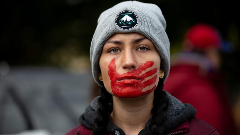 Une jeune femme aux cheveux tressés, vêtue d’une veste bordeaux et coiffée d’un bonnet d’hiver gris, regarde résolument vers l’avant, une empreinte de main rouge peinte sur la bouche et le visage. Visibilité masquée.