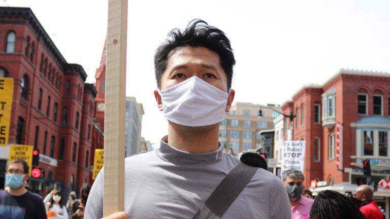 Un homme portant un masque en tissu et tenant une pancarte, marchant avec un groupe de personnes portant des pancartes sur la fin de la violence raciste et du racisme anti-asiatique. Visibilité masquée.