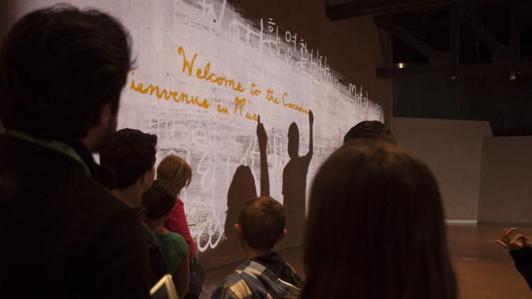 Un groupe de personnes devant un mur avec une projection de deux silhouettes en train d'écrire la phrase « Bienvenue au Musée canadien pour les droits de la personne » en anglais et en français. Visibilité masquée.