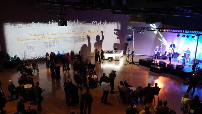 A crowded hall of people watching musicians perform on a brightly lit stage. On the wall in the background, a projected image reads “Welcome to the Canadian Museum for Human Rights.” Visibilité masquée.