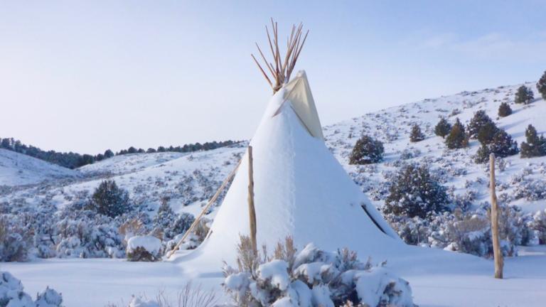 Un tipi recouvert de neige se dresse dans un paysage vallonné parsemé d’arbres et de buissons, sous un ciel bleu. Visibilité masquée.