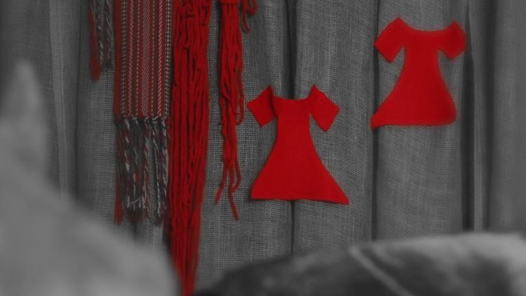 Deux robes en feutre rouge vif sont épinglées sur un fond gris, à côté de laine rouge tressée et d’une ceinture traditionnelle métisse. Visibilité masquée.