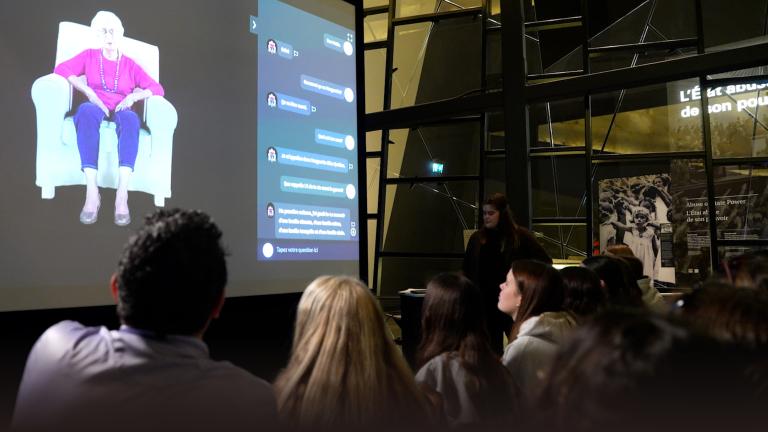 Un auditoire regarde un grand écran de projection sur lequel apparaît une femme âgée vêtue d’un haut violet et d’un pantalon bleu, assise dans un fauteuil blanc, à côté d’une interface interactive. La présentation se déroule dans un espace moderne à l’architecture géométrique en verre, et une personne se tient à droite de l’écran. Visibilité masquée.