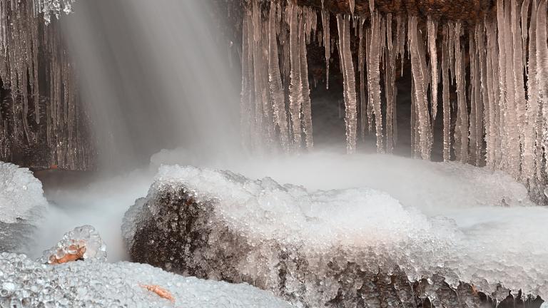 De l’eau est projetée sur des formations rocheuses à partir d’une ouverture invisible située au-dessus, ce qui crée un ensemble spectaculaire de stalactites et de monticules de glace sur les rochers. Visibilité masquée.