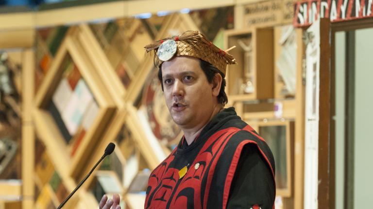 Un homme vêtu d’un costume et d’une coiffe traditionnels autochtones se tient devant une installation d’art faite de panneaux de bois avec des objets encastrés. Visibilité masquée.