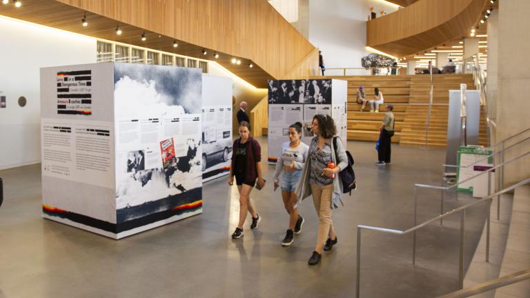 Vue intérieure d’un atrium blanc avec des niveaux en bois au-dessus. Les gens passent devant trois présentoirs d’information autoportants. Les présentoirs contiennent du texte et des images, avec pour toile de fond une architecture en bois et un escalier. Certaines personnes sont en train de discuter, tandis que d’autres interagissent avec l’exposition.