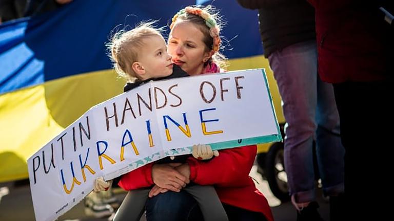 Un enfant tient un panneau de protestation qui dit, en anglais : « Poutine, ne touche pas à l’Ukraine. » L’enfant est assis sur les genoux de la personne qui s'occupe de lui. Derrière eux se trouve un drapeau ukrainien. Visibilité masquée.