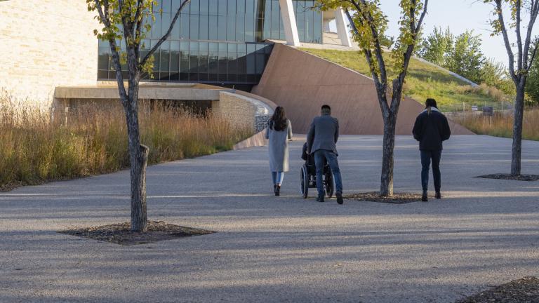 Quatre personnes suivent le chemin menant au Musée canadien pour les droits de la personne par une journée d'automne. Trois d'entre elles sont à pied et une personne est en fauteuil roulant. Visibilité masquée.