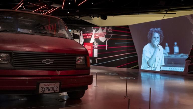 Fourgonnette Chevrolet rouge dans un espace d'exposition avec une vidéo de Rage Against the Machine en arrière-plan. Visibilité masquée.