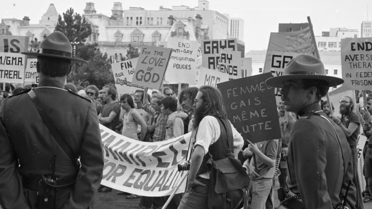 Un grand groupe de protestataires défile dans une ville tandis que deux agents de la GRC en uniforme surveillent la scène. Sur cette image en noir et blanc, les pancartes et les banderoles réclament la fin des pratiques discriminatoires contre les membres de la communauté 2ELGBTQI+. Visibilité masquée.