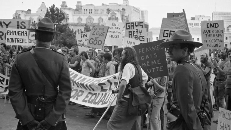 Un grand groupe de protestataires défile dans une ville tandis que deux agents de la GRC en uniforme surveillent la scène. Sur cette image en noir et blanc, les pancartes et les banderoles réclament la fin des pratiques discriminatoires contre les membres de la communauté 2ELGBTQI+. Visibilité masquée.