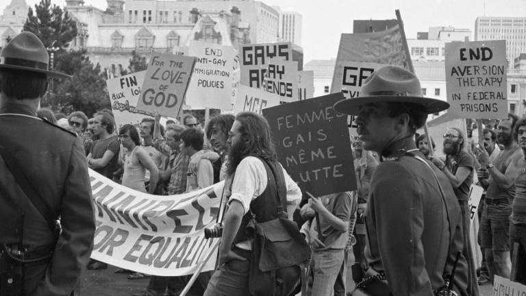 Un grand groupe de protestataires défile dans une ville tandis que deux agents de la GRC en uniforme surveillent la scène. Sur cette image en noir et blanc, les pancartes et les banderoles réclament la fin des pratiques discriminatoires contre les membres de la communauté 2ELGBTQI+. Visibilité masquée.