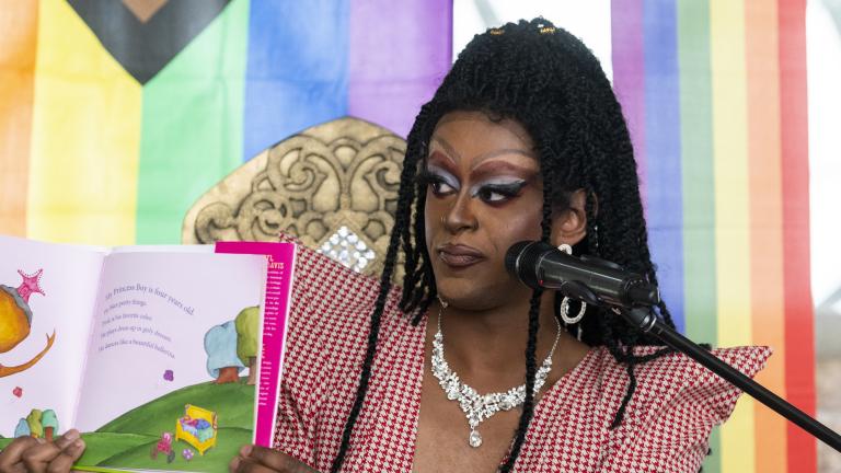 Une drag queen très maquillée et coiffée d’une perruque composée de longues et minuscules tresses brandit un livre pour enfants pour le montrer à un public. La toile de fond derrière elle est un arc-en-ciel pastel. Visibilité masquée.