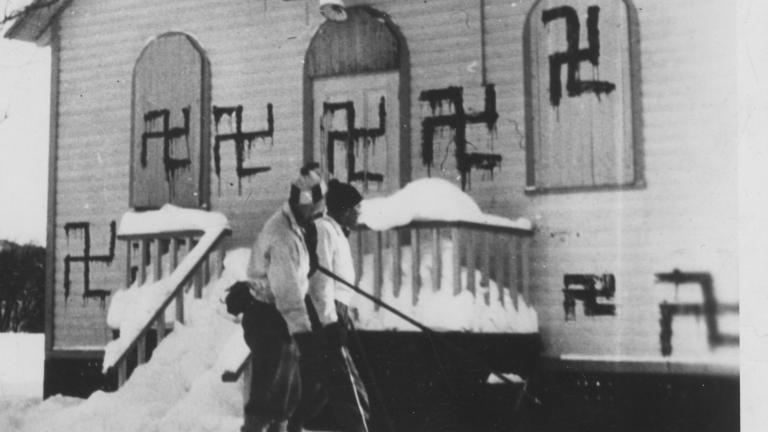 Deux personnes jouent au hockey devant une synagogue où plusieurs croix gammées ont été peintes, Sainte-Marguerite, Québec, 1938. Visibilité masquée.