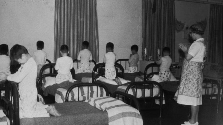 Un groupe de garçons en pyjama est agenouillé sur des lits simples, la tête baissée et les mains jointes comme pour prier. Une femme se tient dans la pièce, les mains jointes de la même manière. Visibilité masquée.