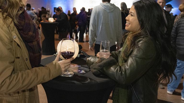 Deux femmes souriantes boivent du vin dans une grande salle remplie de gens. Visibilité masquée.