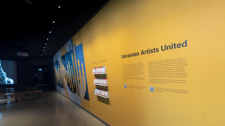Un mur jaune avec les mots "Ukrainian Artists United" et un graphique dynamique bleu et jaune se trouve à droite de l'image. Sur le côté gauche, une image est pr ojectée sur un écran montrant une personne avec des écouteurs et la bouche ouverte, comme si elle chantait. Des bancs pour s'asseoir sont placés devant l'écran. Visibilité masquée.