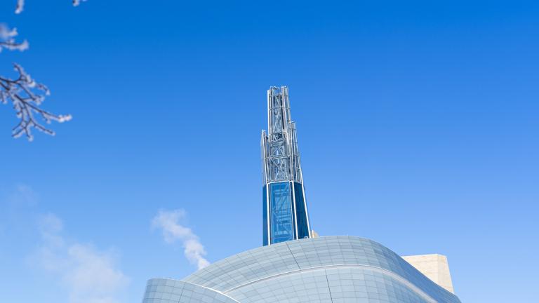 Une scène hivernale en plein air avec une vue extérieure du Musée canadien pour les droits de la personne, d’autres bâtiments au loin et des arbres recouverts de givre. Visibilité masquée.