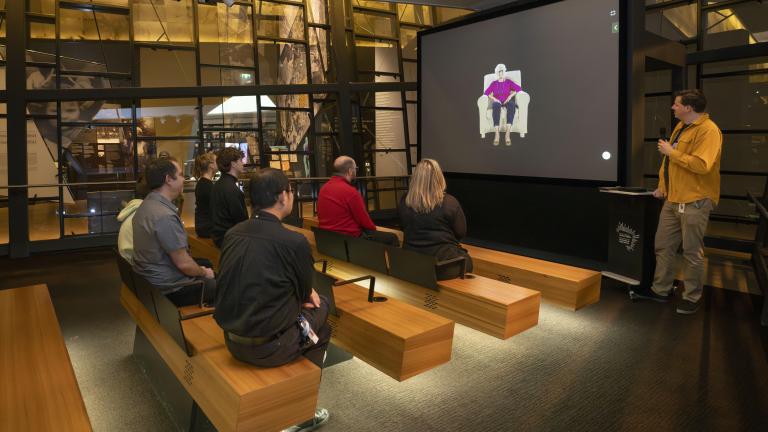 Un petit groupe de personnes assises sur des bancs en bois dans un théâtre fait face à un écran montrant une femme âgée assise dans un fauteuil. Un homme vêtu d’une chemise jaune se tient à droite de l’écran et tient un microphone. Visibilité masquée.