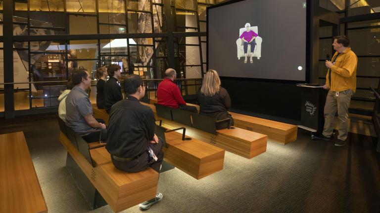 Un petit groupe de personnes assises sur des bancs en bois dans un théâtre fait face à un écran montrant une femme âgée assise dans un fauteuil. Un homme vêtu d’une chemise jaune se tient à droite de l’écran et tient un microphone. Visibilité masquée.