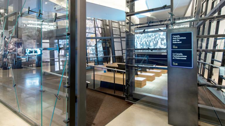 Des parois de verre inégales entourent une salle de projection avec un écran et 4 bancs en face. À l’extérieur de l’entrée de la salle, un panneau indique le film sélectionné. Visibilité masquée.