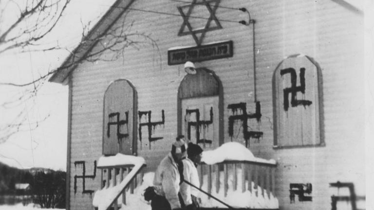 Deux personnes jouent au hockey devant une synagogue où plusieurs croix gammées ont été peintes, Sainte-Marguerite, Québec, 1938. Visibilité masquée.
