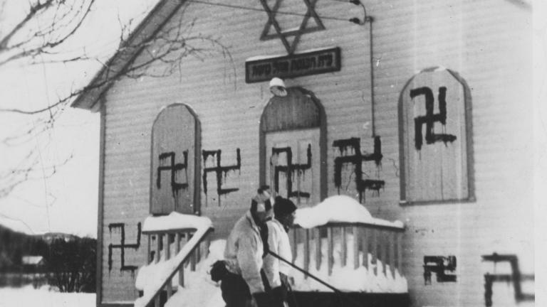Deux personnes jouent au hockey devant une synagogue où plusieurs croix gammées ont été peintes, Sainte-Marguerite, Québec, 1938. Visibilité masquée.