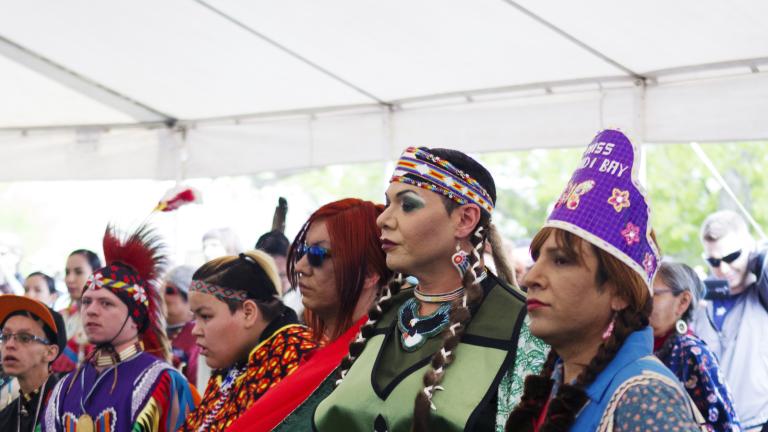 Un groupe de personnes portant des vêtements autochtones colorés sont alignées à l’intérieur d’une tente sur de l’herbe et dansent.