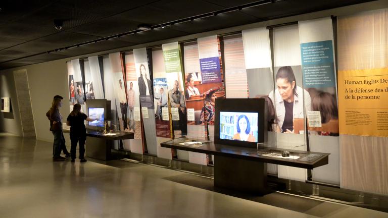 Deux personnes interagissent avec l’un de deux écrans vidéo dans une galerie de musée. Il y a des images de gens et des blocs de texte en couleur sur le mur.