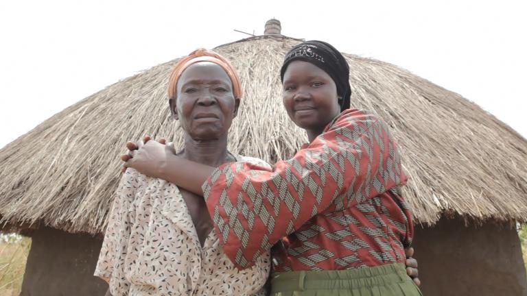 Deux femmes debout devant une hutte avec un toit de paille, regardent la caméra. La femme plus à droite sourit en serrant l’autre femme dans ses bras.