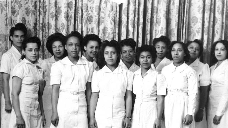Une image en noir et blance de Viola Desmond avec 11 autres femmes. Elles portent toutes des robes d’un blanc uni et se tiennent debout en deux rangées. Elles regardent vers l’objectif. 