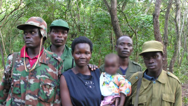 Une femme tenant un bébé, tandis que quatre hommes en tenue militaire se tiennent à côté et derrière elle. Ils sont tous debout devant une forêt et posent pour la photo.