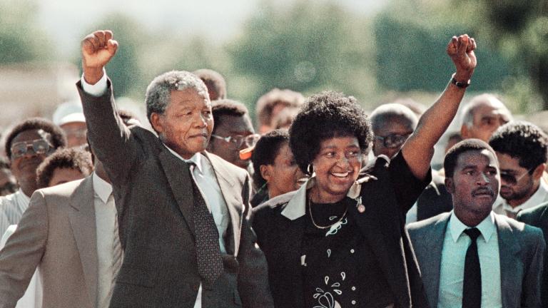 Nelson Mandela, an older black man with graying hair, forms a raised fist with his right hand and holds it above his head. With his left, he is holding hands with Winnie Mandela, a black woman with dark hair. She is smiling and has formed a raised fist with her left hand, mirroring Nelson Mandela’s raised fist. A large crowd is gathered behind them.