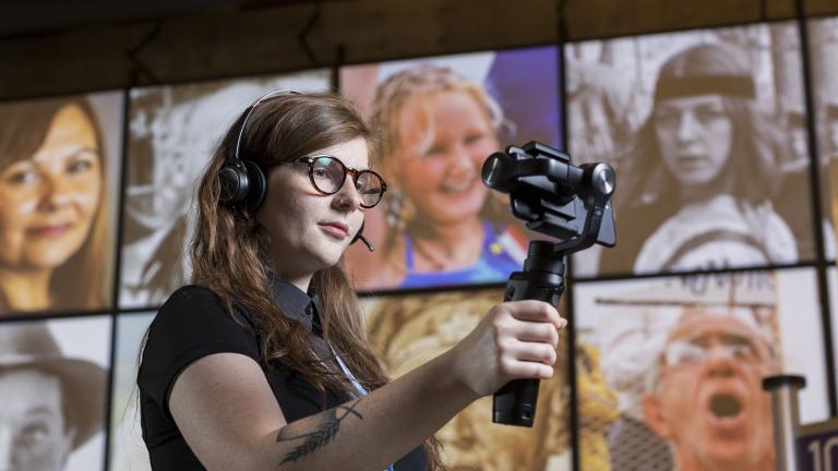 Une femme portant des écouteurs, tenant un appareil mobile fixé sur une poignée, se tient dans une galerie du Musée.
