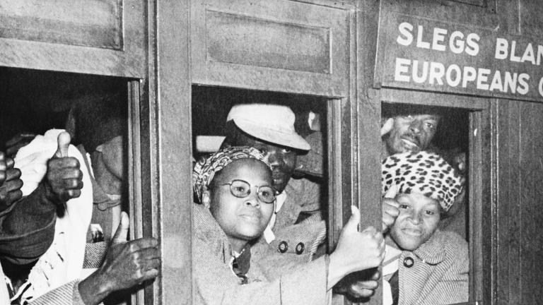 Une image en noir et blanc d’un groupe de femmes et d’hommes noirs à l’intérieur d’un wagon qui regardent à l’extérieur par les fenêtres. Plusieurs ont la main levée et tiennent le pouce en l’air. Au-dessus d’une fenêtre, il est écrit que le wagon est réservé à la population européenne.