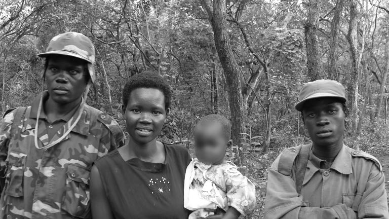 Une photo en noir et blanc d'une femme tenant un bébé dans ses bras, tandis que deux hommes en tenue militaire se tiennent à ses côtés. Ils sont debout devant une forêt et posent pour la photo.