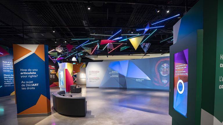 Une galerie de musée où on voit des oeuvres d'art projetées sur de grands écrans et formes géométriques colorées.