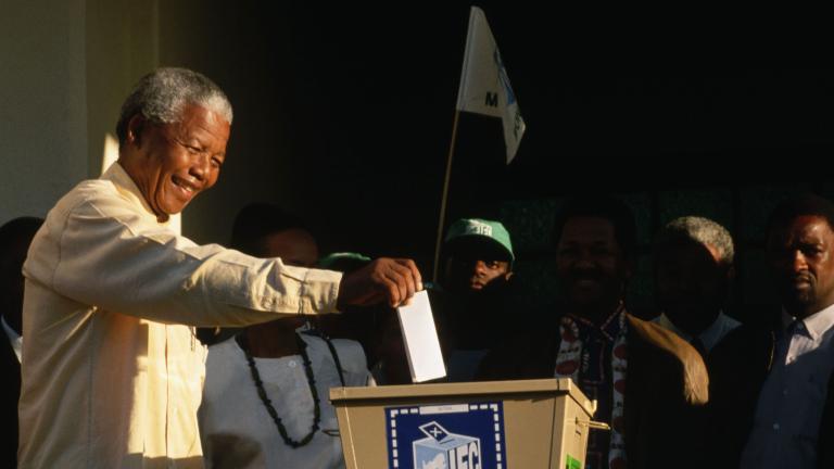 Un Nelson Mandela âgé sourit en tendant le bras droit pour déposer son bulletin de vote dans une grande boîte de scrutin en métal.