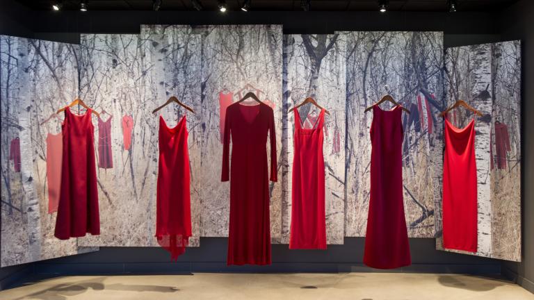 Six robes rouges sont suspendues à des cintres devant une toile de fond. Cette toile de fond montre l’image d’une forêt de bouleaux dans laquelle d’autres robes rouges sont suspendues.