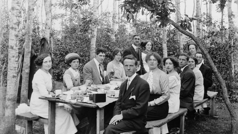 Un groupe d’hommes et de femmes bien habillés assis à une table de pique-nique avec des arbres en arrière-plan. 