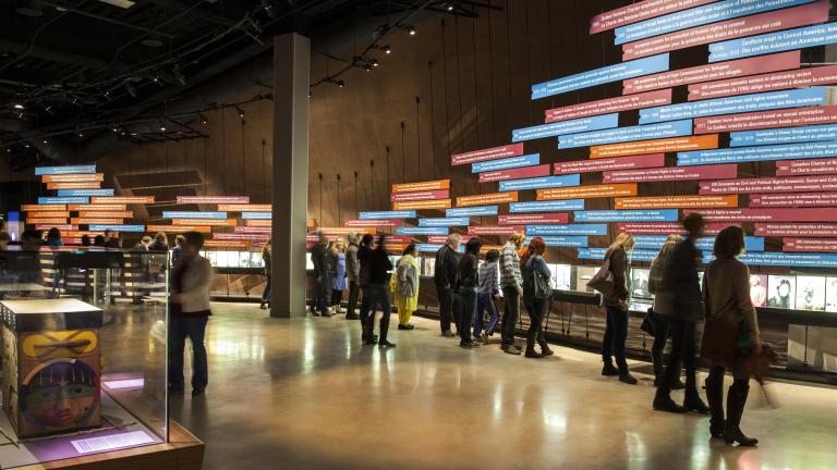 Une grande foule dans une galerie du Musée. La plupart des gens regardent une exposition sur le mur de la galerie. L'exposition comprend une série d'images et de longs panneaux rectangulaires de texte, placés horizontalement.