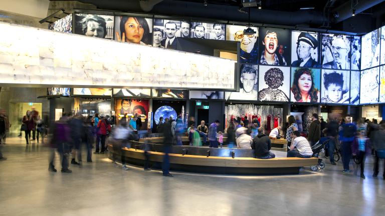 Une grande foule dans une galerie de musée. Il y a un banc courbe au centre de la salle. Dans l’espace, on voit une série de grandes images placées au-dessus d’alcôves qui contiennent chacune une exposition différente.
