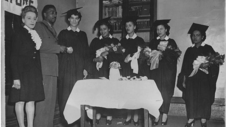 Une image en noir et blanc de Viola Desmond debout à côté d’un homme en uniforme. Debout à leur gauche, cinq femmes portent des coiffes et des robes de finissante et tiennent des fleurs. Une femme accepte un parchemin roulé de l’homme en uniforme.