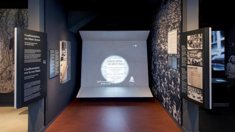 Une exposition muséale comprenant des photos, du texte et un grand écran vidéo avec des sous-titres et une interprétation en langue des signes.