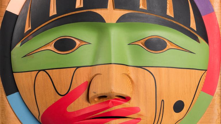Une boîte en bois, sur laquelle est sculpté un visage dont la bouche est recouverte d’une main peinte en rouge.