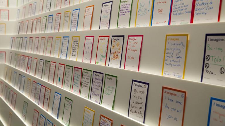 Des cartes avec l’expression « J’imagine… » en français et en anglais sont placées le long d’un mur blanc sur plusieurs rangées de présentoirs. Des personnes en visite au Musée ont dessiné ou écrit sur chaque carte avec des marqueurs.
