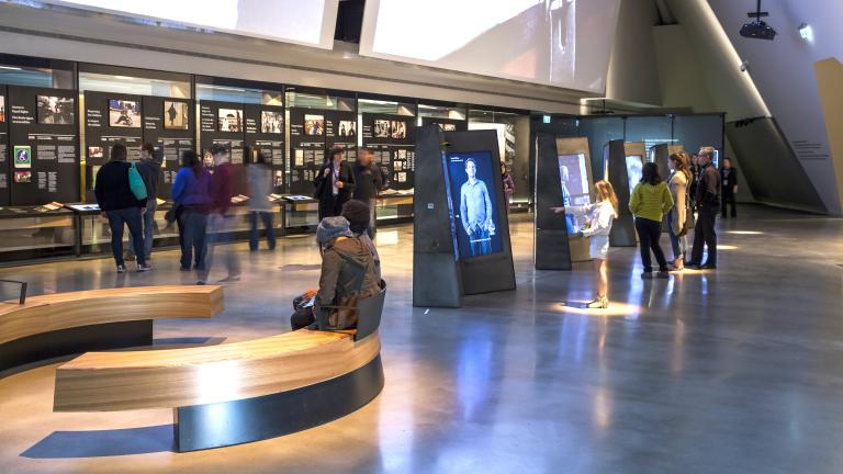 Des gens explorent une galerie contenant des vidéos interactives verticales au centre de la salle et des présentoirs avec texte et images qui longent le mur arrière.